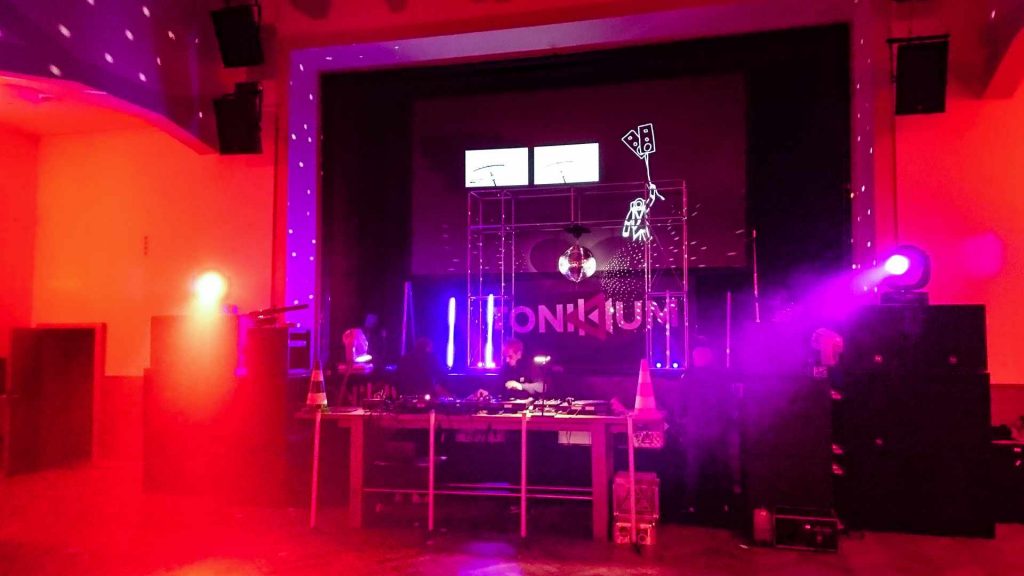 DJ am Mischpult + Techniker am Aufbau der Bühne. Moving- Heads,Projektionen, sowie Soundanlagen sind zu sehen.